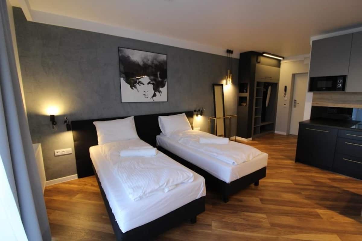 A modern bedroom with two single beds, a gray wall with a black and white artwork, a wooden floor, and a kitchenette in the background.