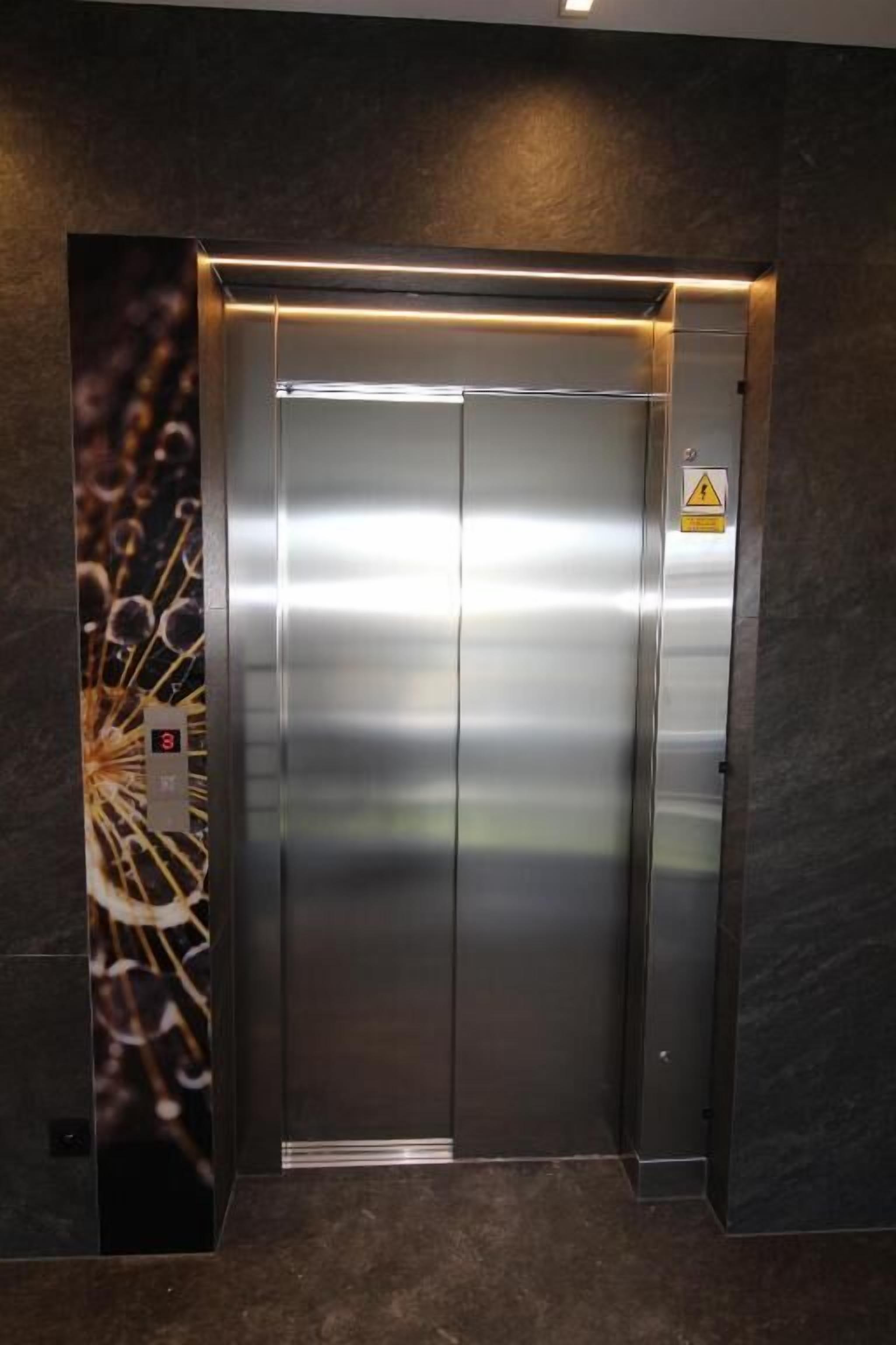 Un ascensor modern amb acabat metàl·lic, un panell de control a la dreta i un senyal de precaució sobre la porta.