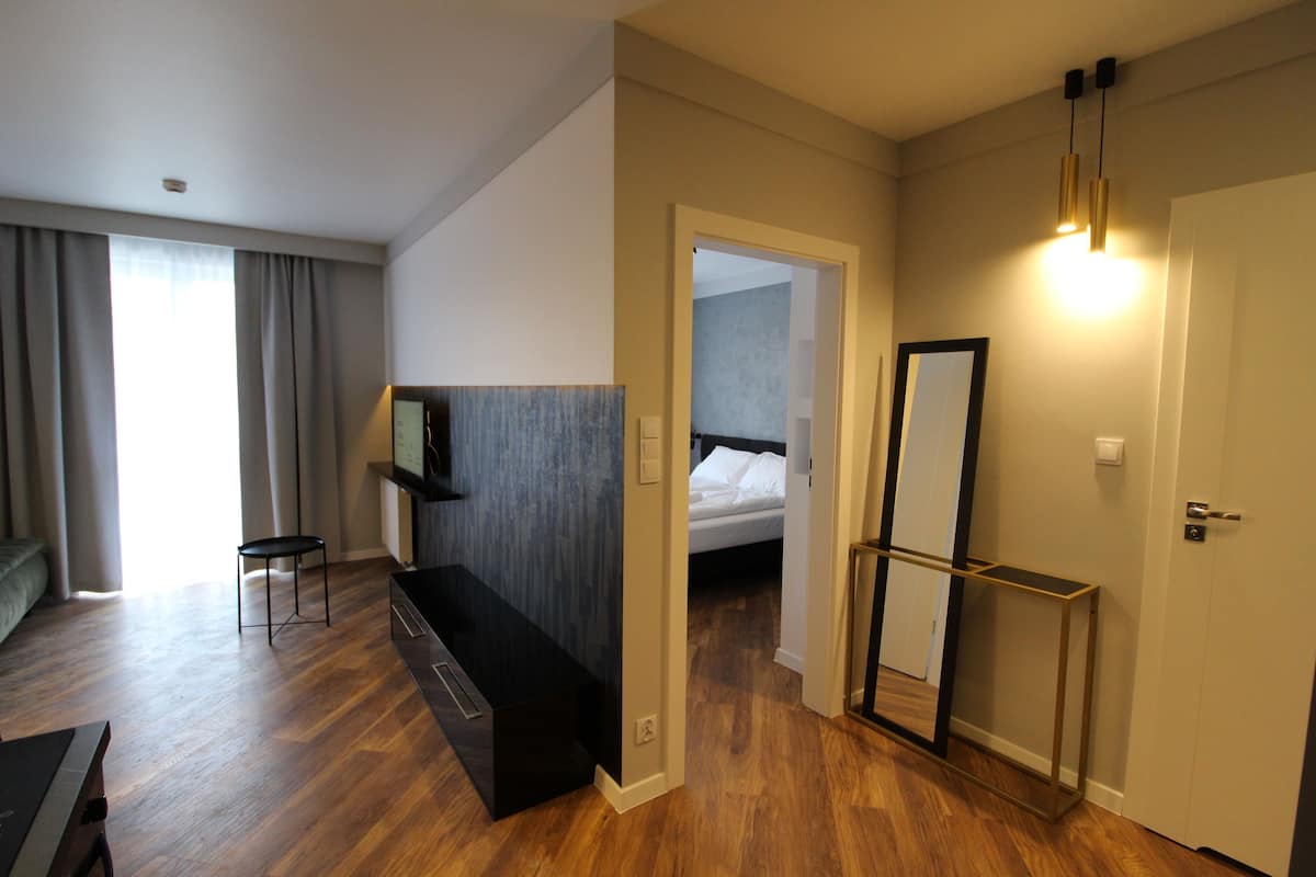 A modern hotel room with a wooden floor, a bed, a desk, a chair, a mirror, and a door.