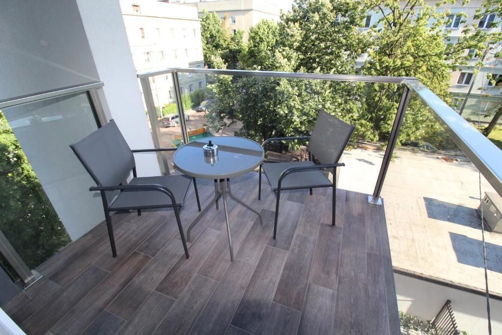A balcony with a round table and two chairs, a metal railing, and a view of a residential area with trees and buildings.