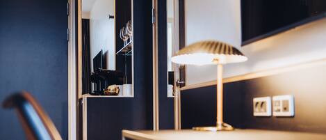 A hotel room with a desk, chair, lamp, and a mirror reflecting a shelf with glasses and a coffee machine.