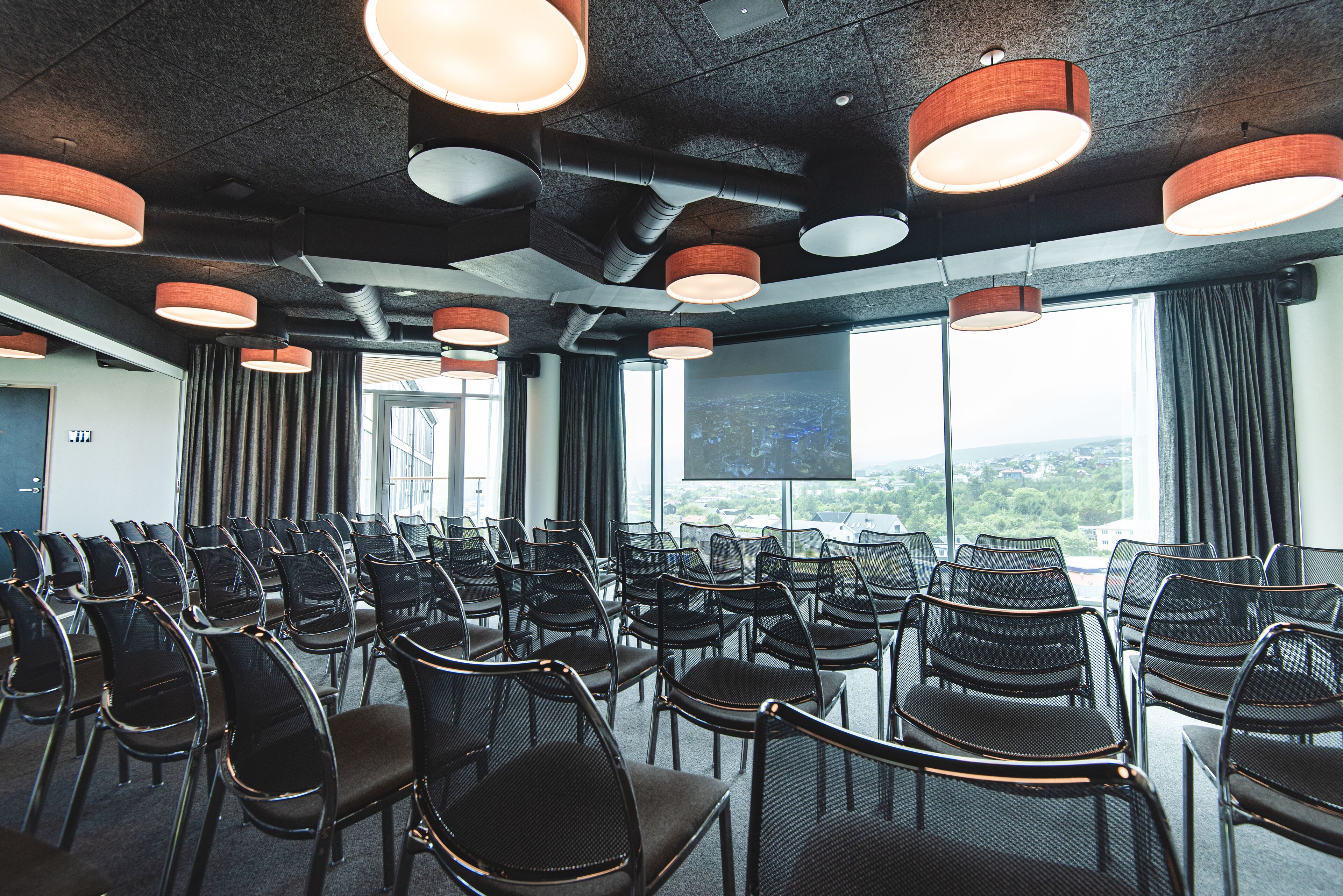 Un salón de conferencias con filas de sillas, ventanas amplias e iluminación colgante moderna.