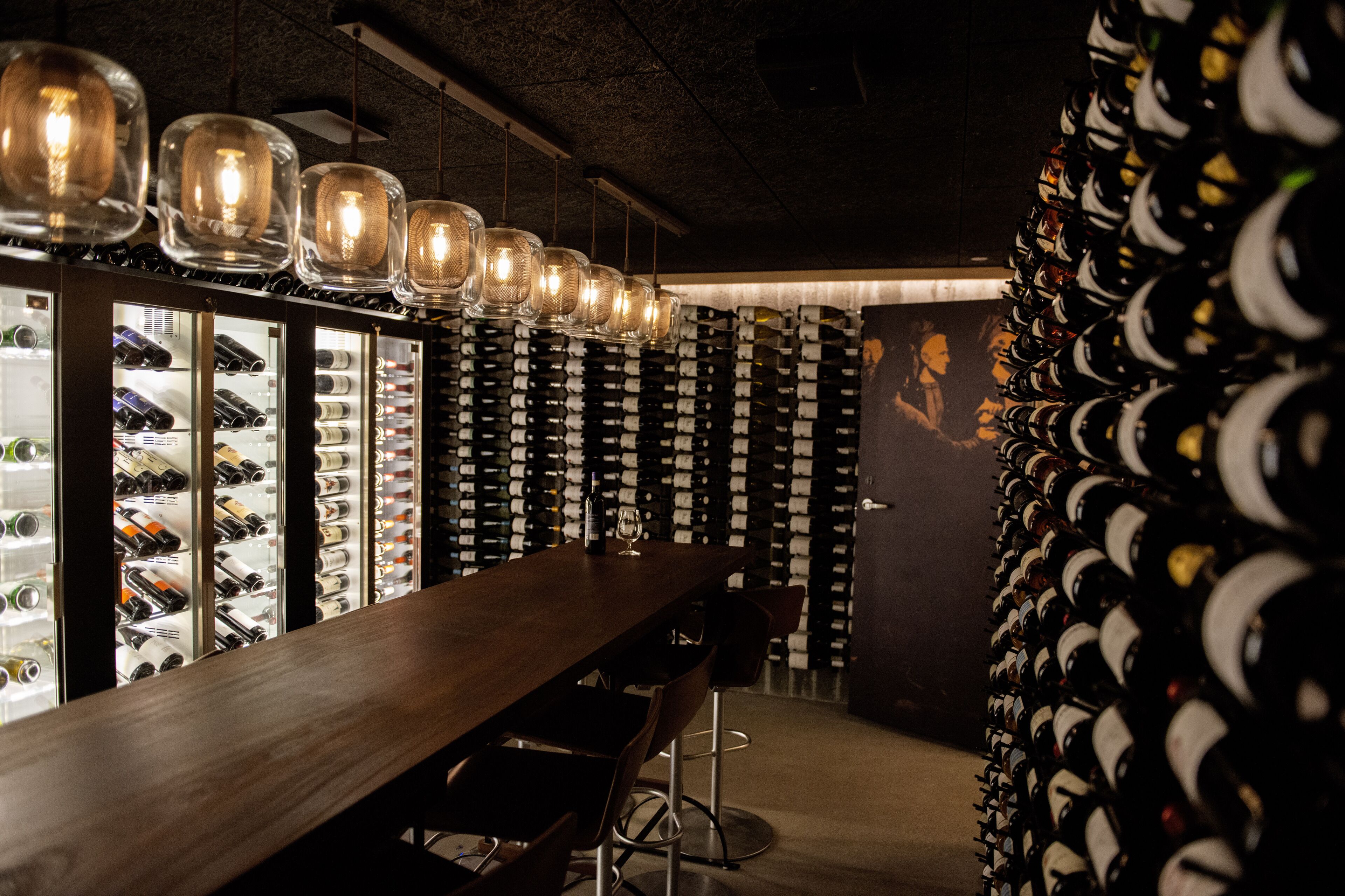 Un espace bar doté d’un long comptoir en bois, de présentoirs à bouteilles et de luminaires suspendus.
