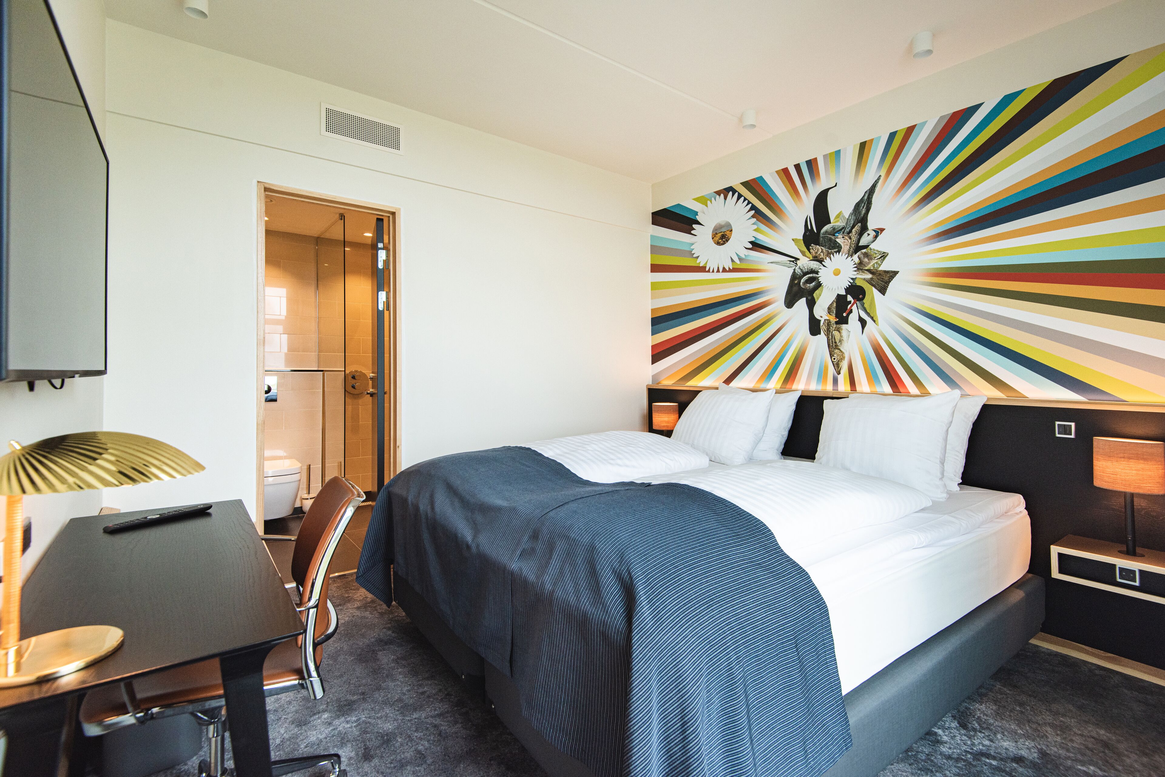 Una habitación de hotel con una cama, un escritorio, una silla, una lámpara y una decoración de pared colorida.