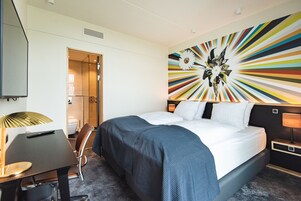 Una habitación de hotel con una cama, un escritorio, una silla, una lámpara y una decoración de pared colorida.