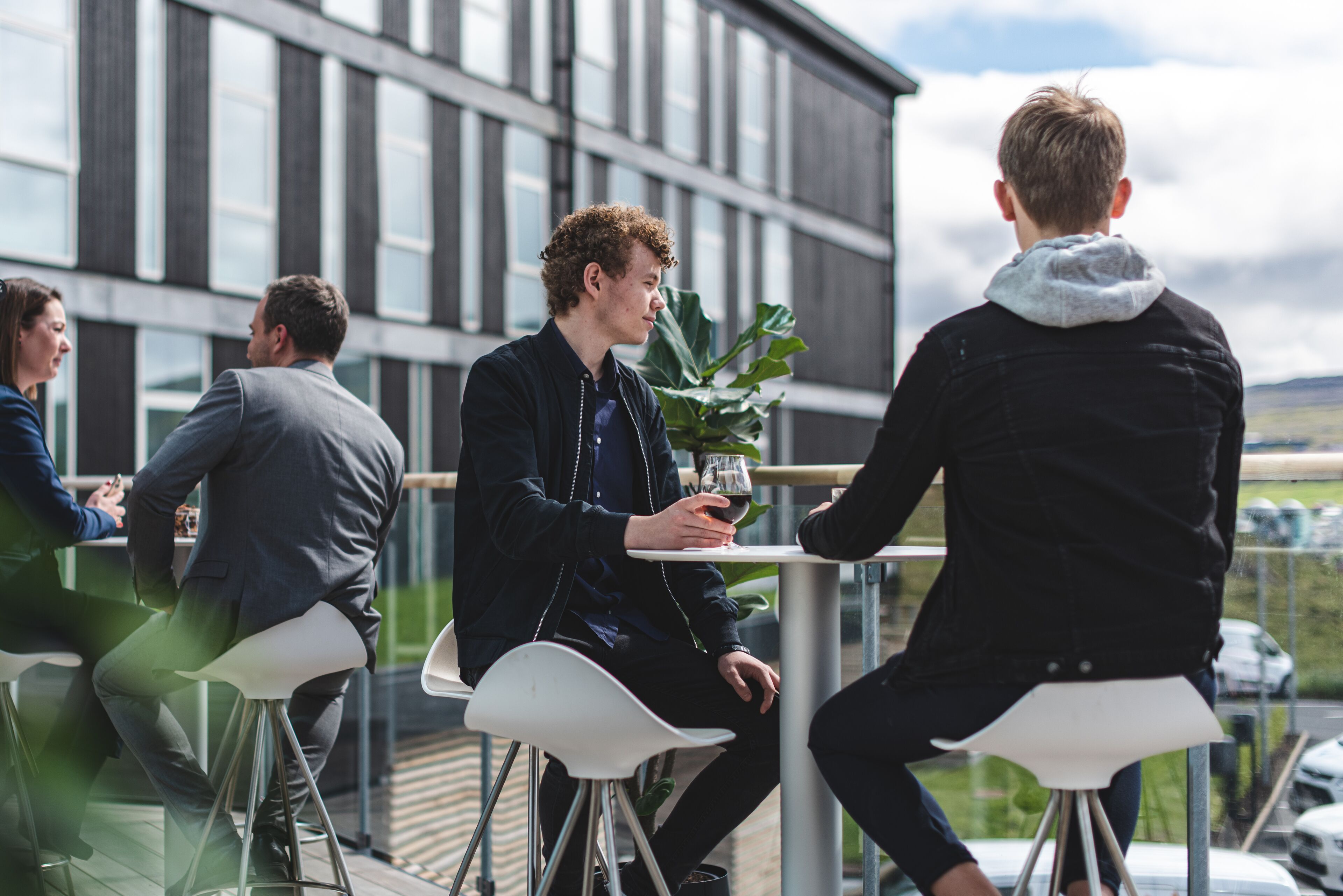 Un groupe de personnes est assis à des tables à l’extérieur, avec un bâtiment moderne en arrière-plan.