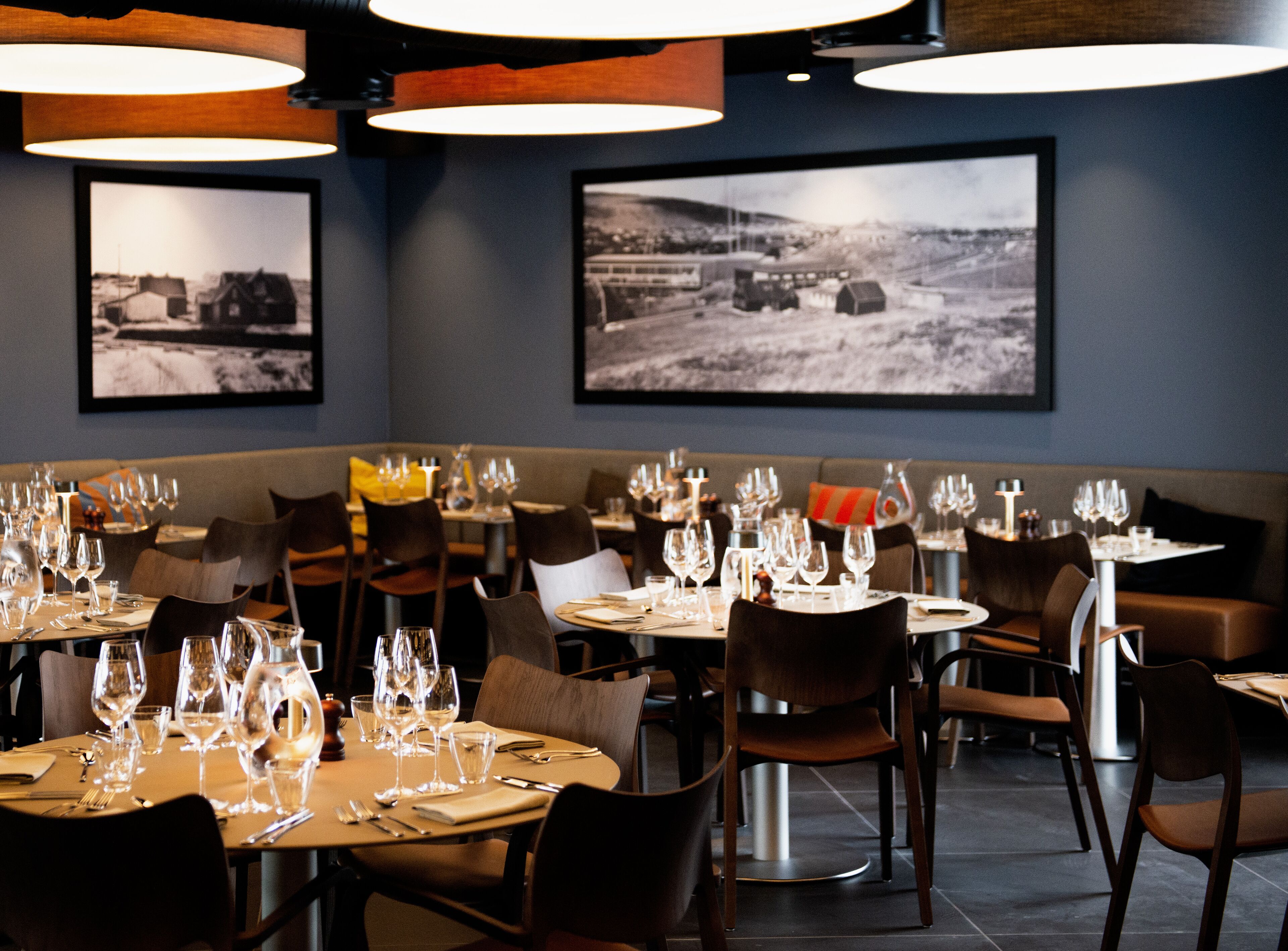 A restaurant with multiple tables set for dining, featuring a modern interior with pendant lighting and framed black-and-white photographs on the walls.