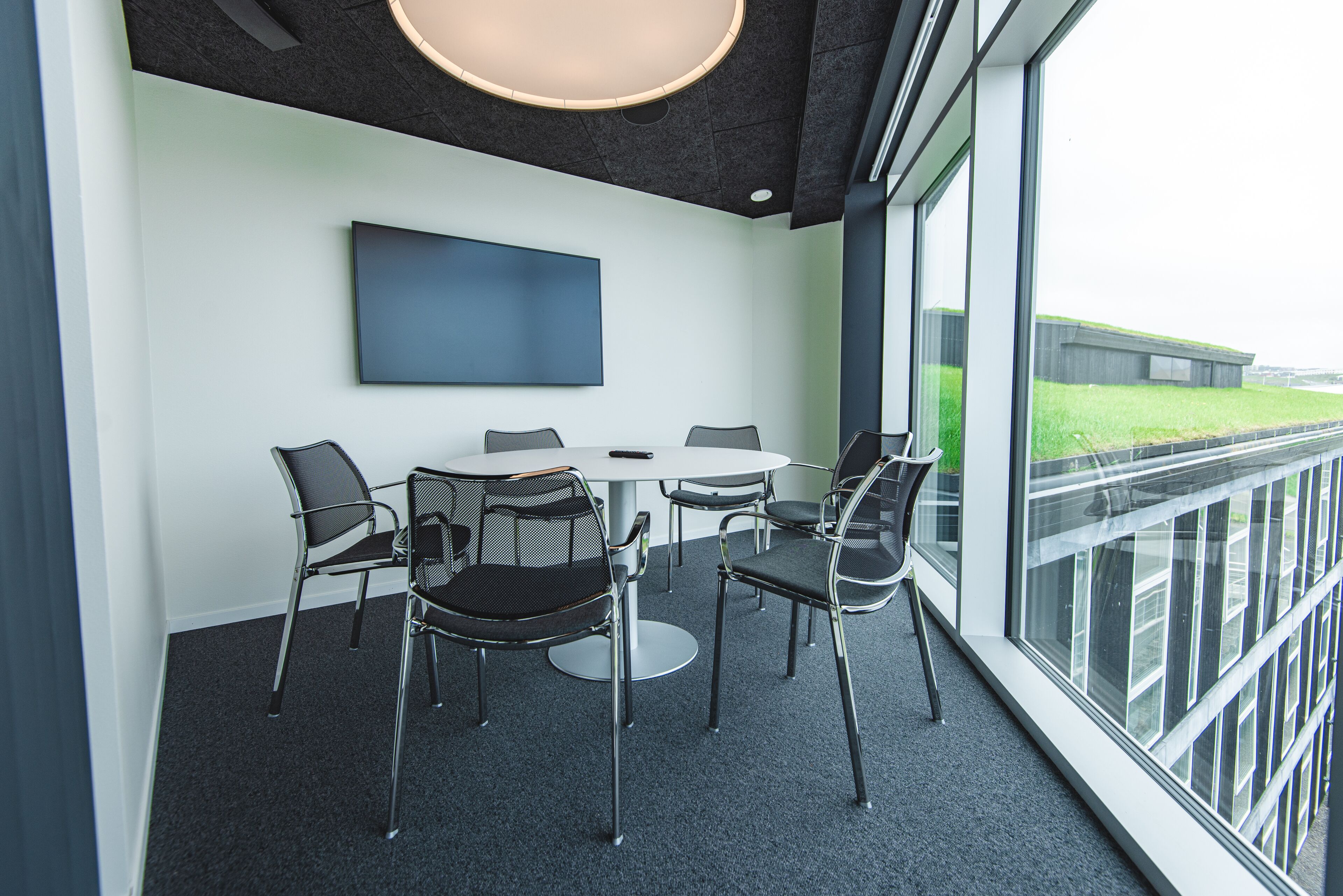Una sala de reuniones moderna con una mesa redonda, cuatro sillas, un televisor de pantalla plana y grandes ventanales que ofrecen vistas al exterior.