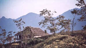 A rustic thatched-roof hut surrounded by wildflowers and trees, with mountains in the background. - Greenland Sa Pa Hotel by DLMH (Sa Pa)
