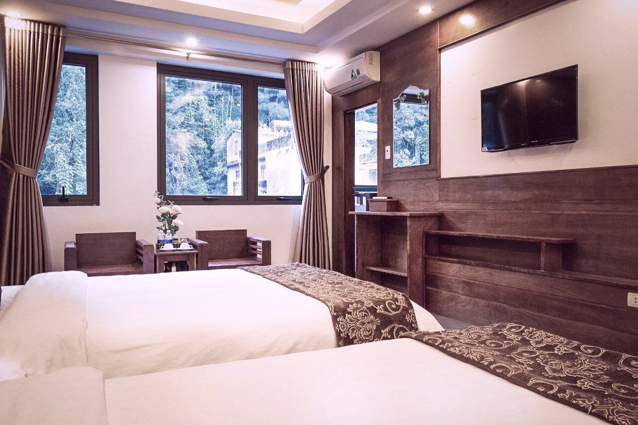 A hotel room with two beds, a TV mounted on the wall, a small table with a vase, and a view of trees through the windows.