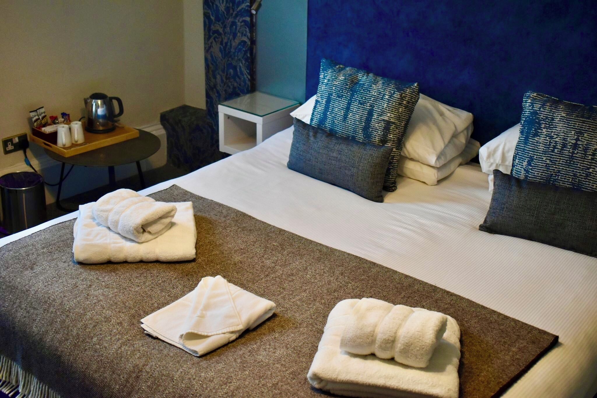 A hotel room with a bed, pillows, a tray with toiletries, and a small table with a kettle.