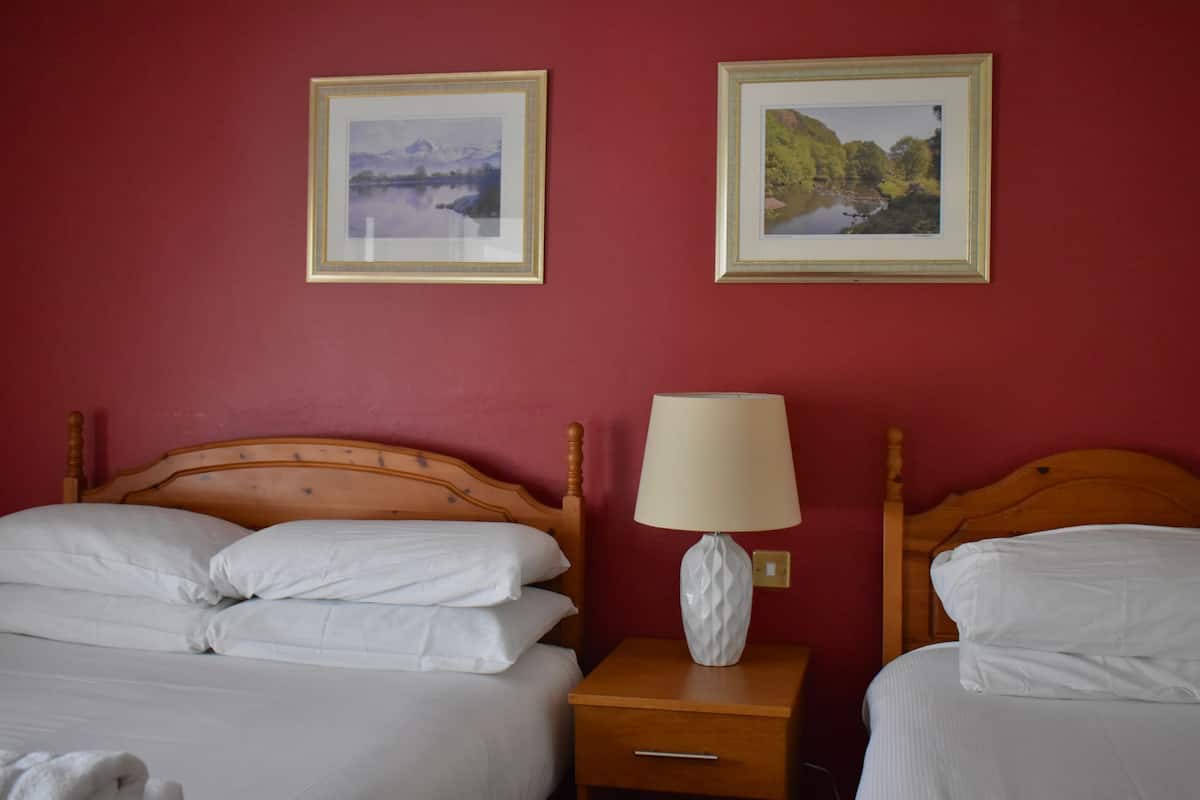 A hotel room with two beds, a wooden headboard, a nightstand with a lamp, and two framed pictures on the wall.