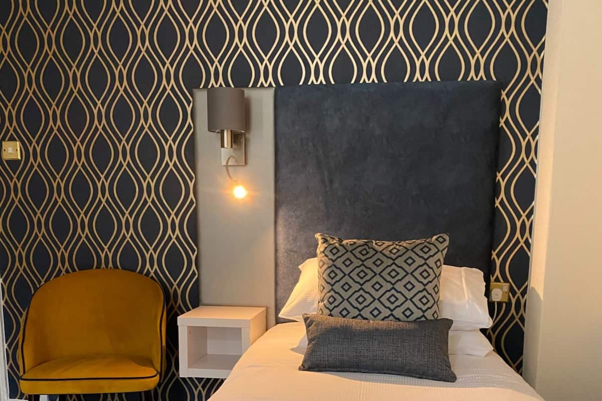A neatly arranged bedroom with a patterned wall, a single bed with pillows, a bedside table, and a chair.