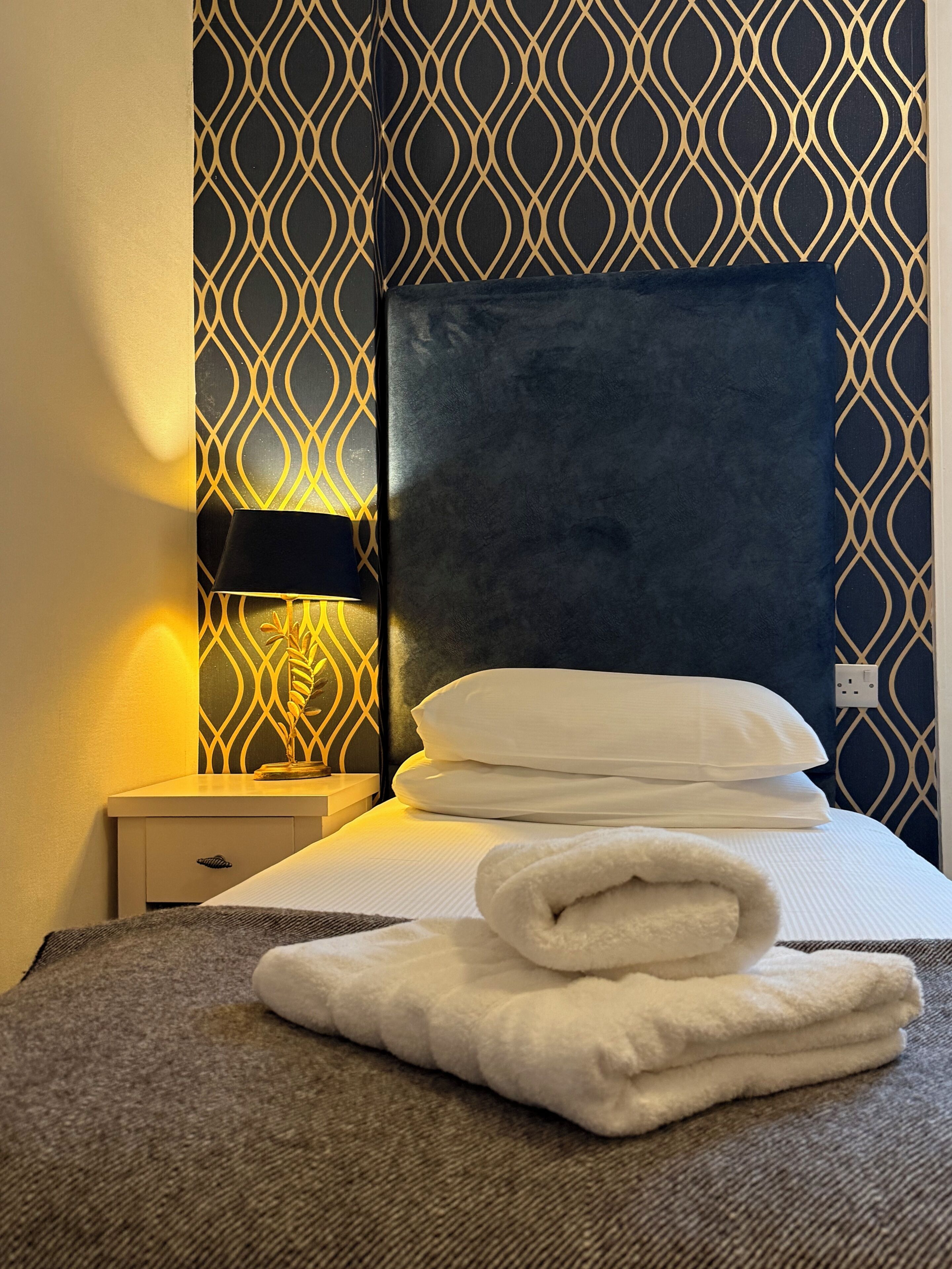 A neatly made bed with white linens and a folded towel, a dark headboard, and a patterned wall.