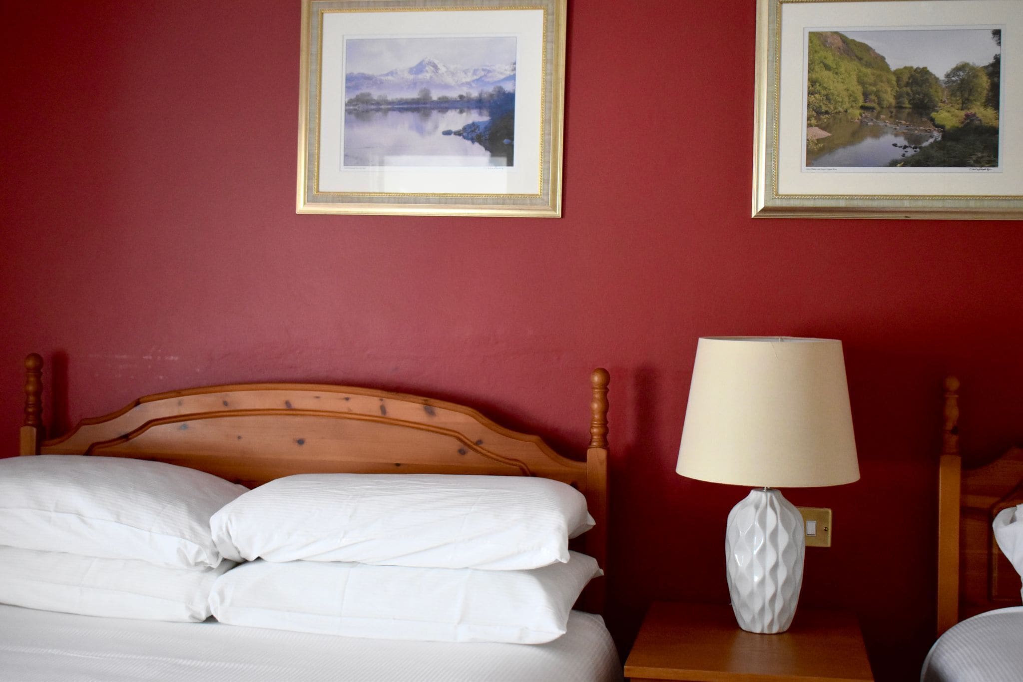 A hotel room with a wooden bed, white bedding, a nightstand with a lamp, and two framed pictures on the wall.