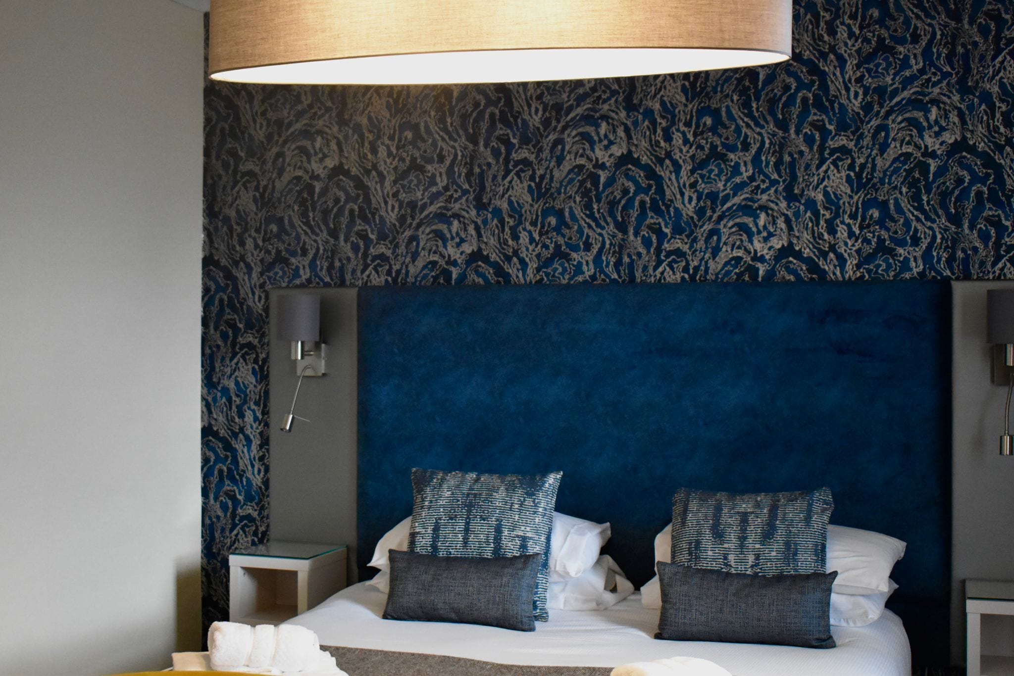 A hotel room with a large bed, two pillows, a bedside table, and a wall-mounted lamp.