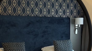 Ein Hotelzimmer mit einem großen Bett, einem Kopfende mit geometrischem Muster, zwei gemusterten Kissen, einer Nachttischlampe und einem runden Spiegel.