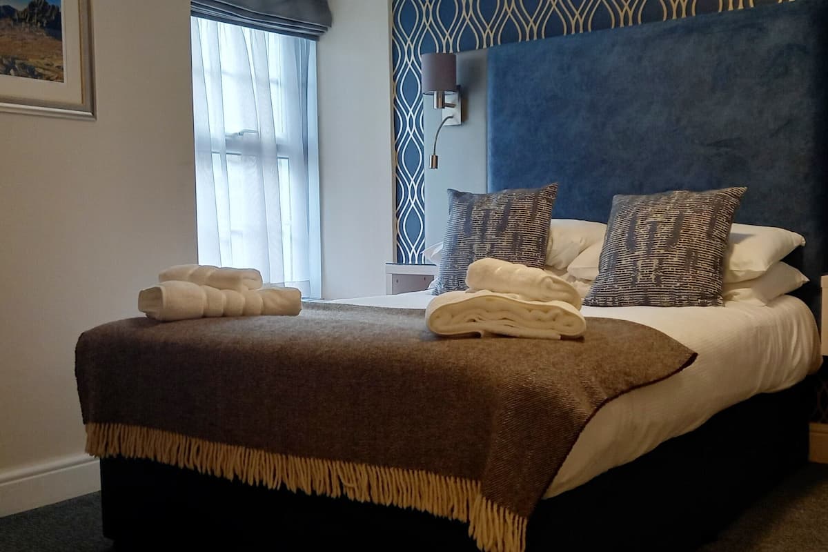 A neatly made bed with a brown throw blanket and two pillows, a bedside table with a lamp, and a framed picture on the wall.
