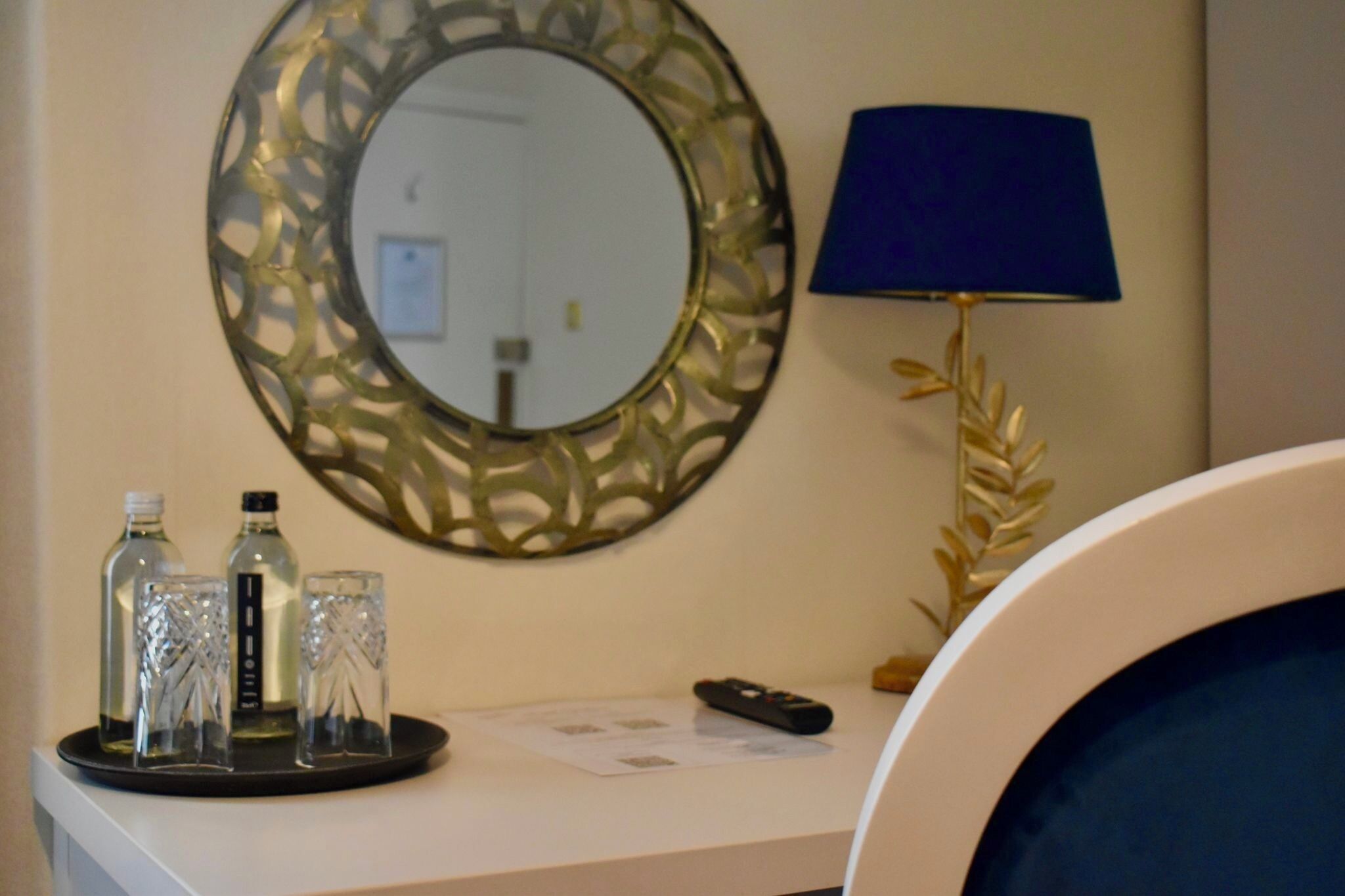 A hotel room with a round mirror, a blue lamp, and a tray with three glasses and two bottles.