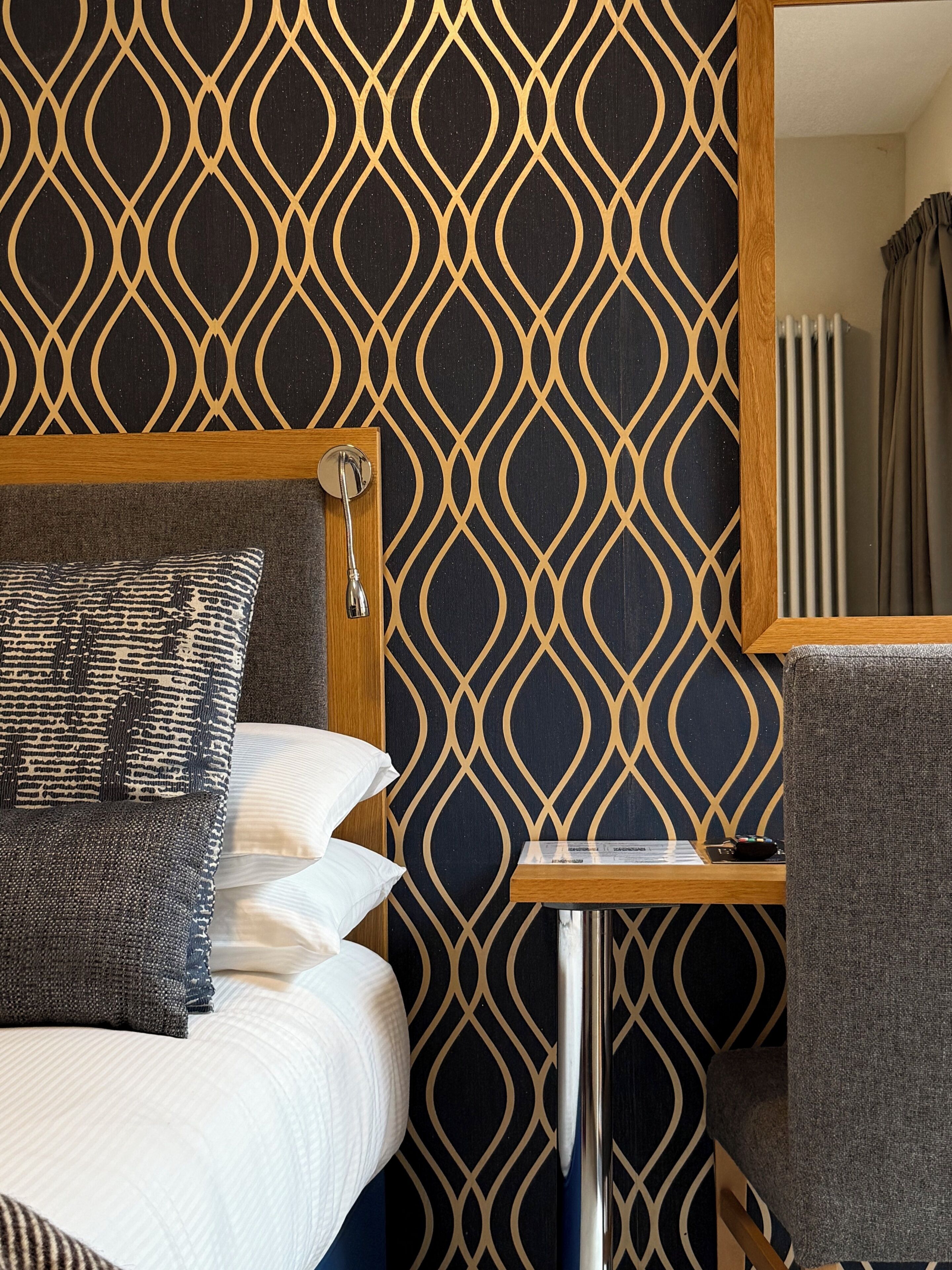 A hotel room with a patterned wallpaper, a bed with pillows, a bedside table, and a chair.
