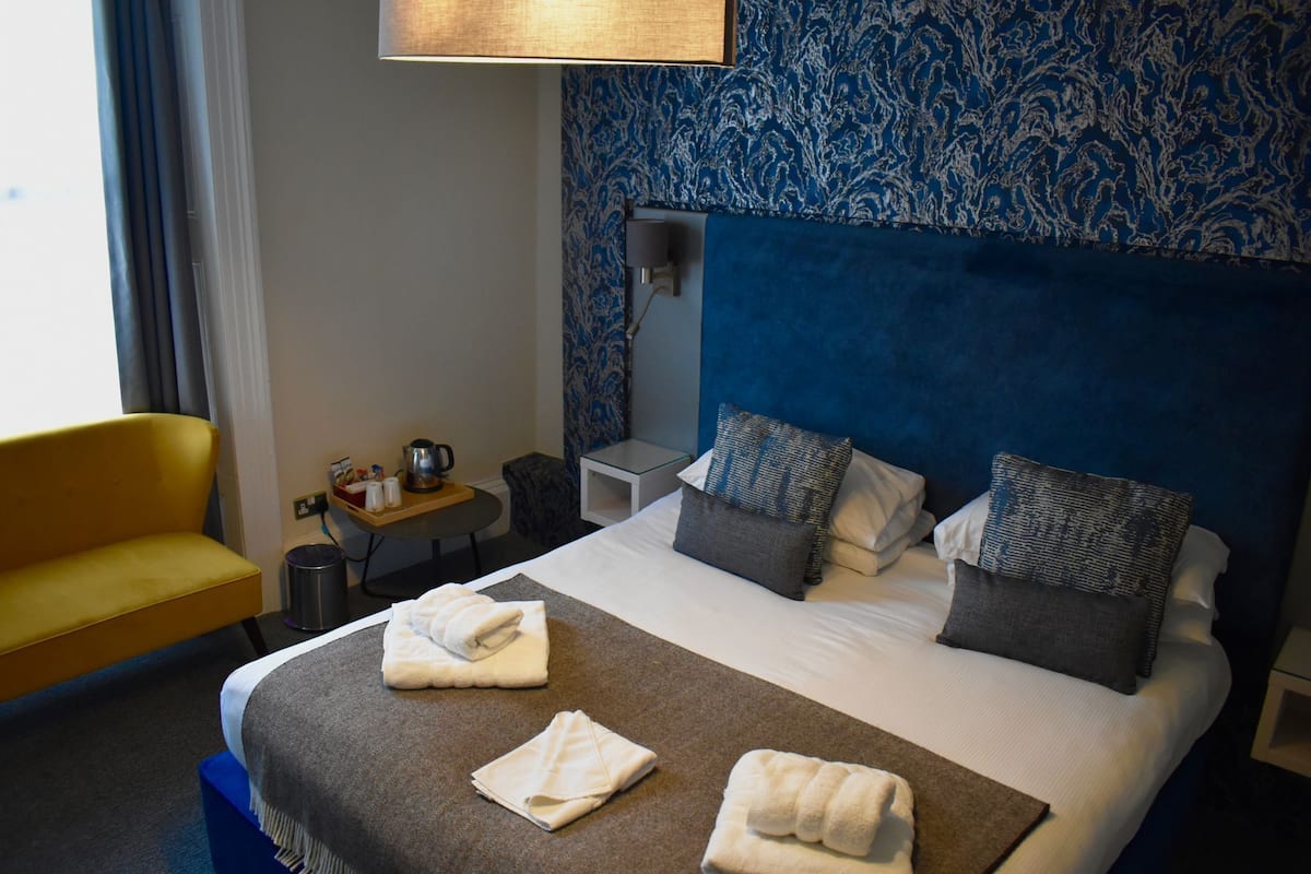 A hotel room with a large bed, two pillows, a bedside table, a lamp, a chair, and a blue patterned wall.