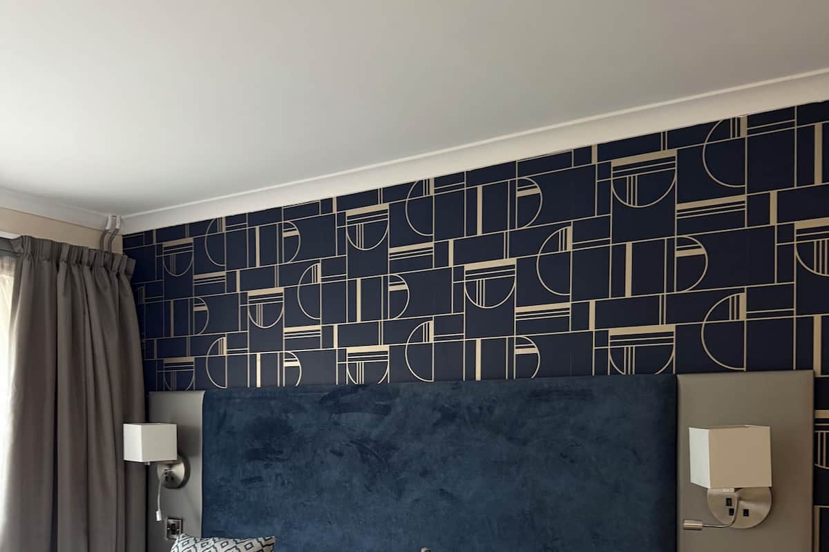 A hotel room with a large bed, a bedside table, a nightstand, a wall-mounted lamp, and a patterned wall.