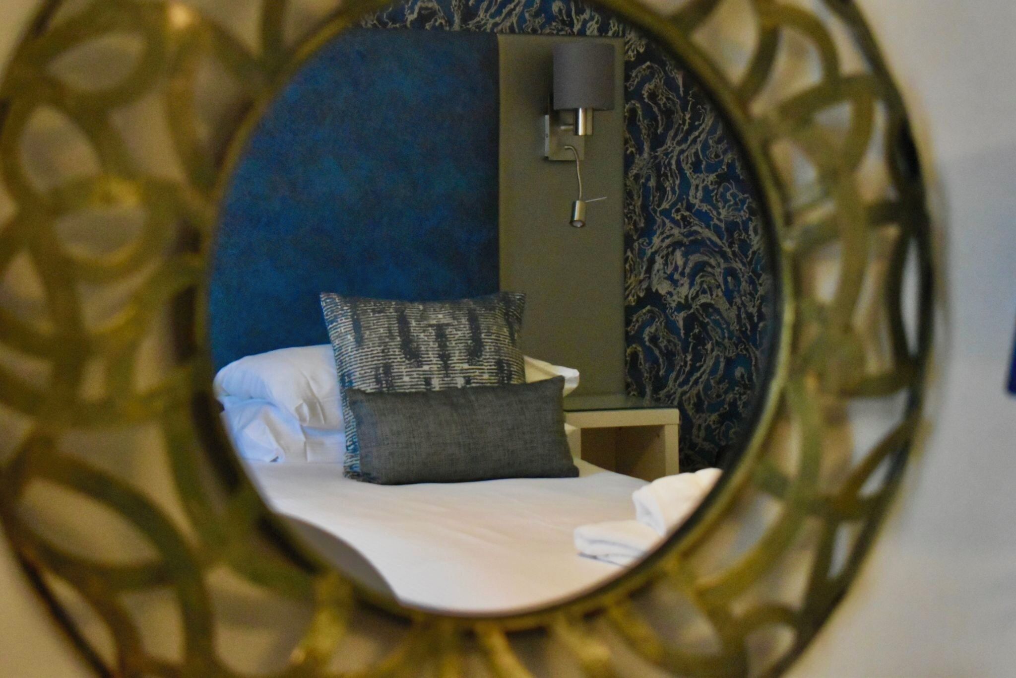 Un lit avec des oreillers, une table de chevet et une lampe fixée au mur, dans une pièce dont le papier peint est à motifs.