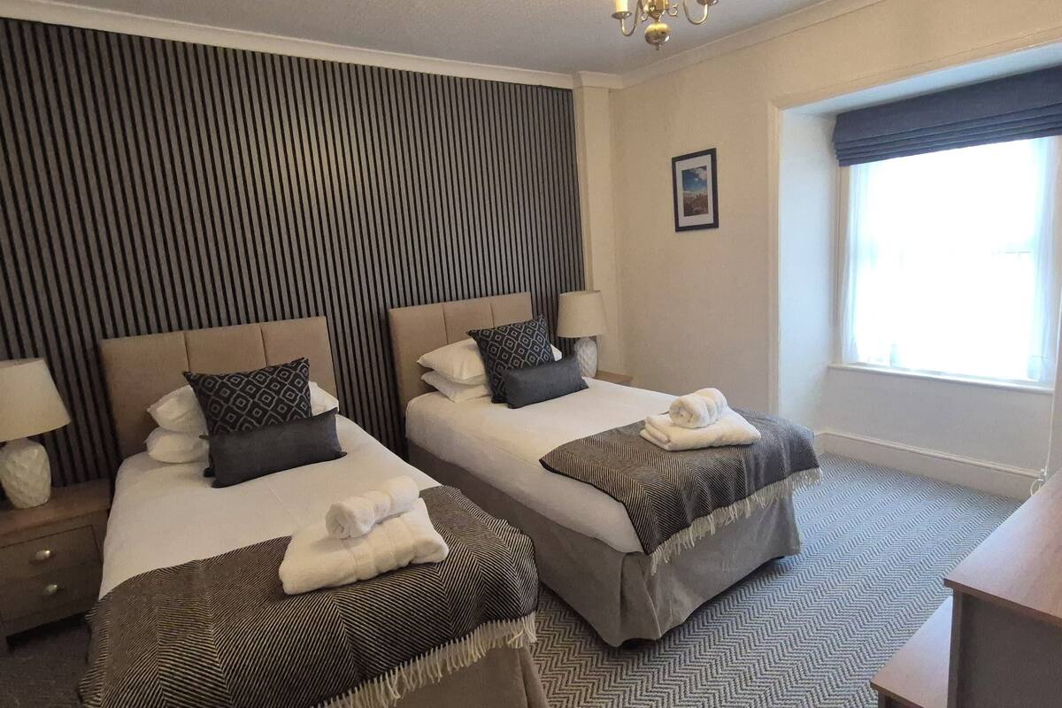 A hotel room with two beds, a nightstand with a lamp, a window with curtains, and a picture on the wall.