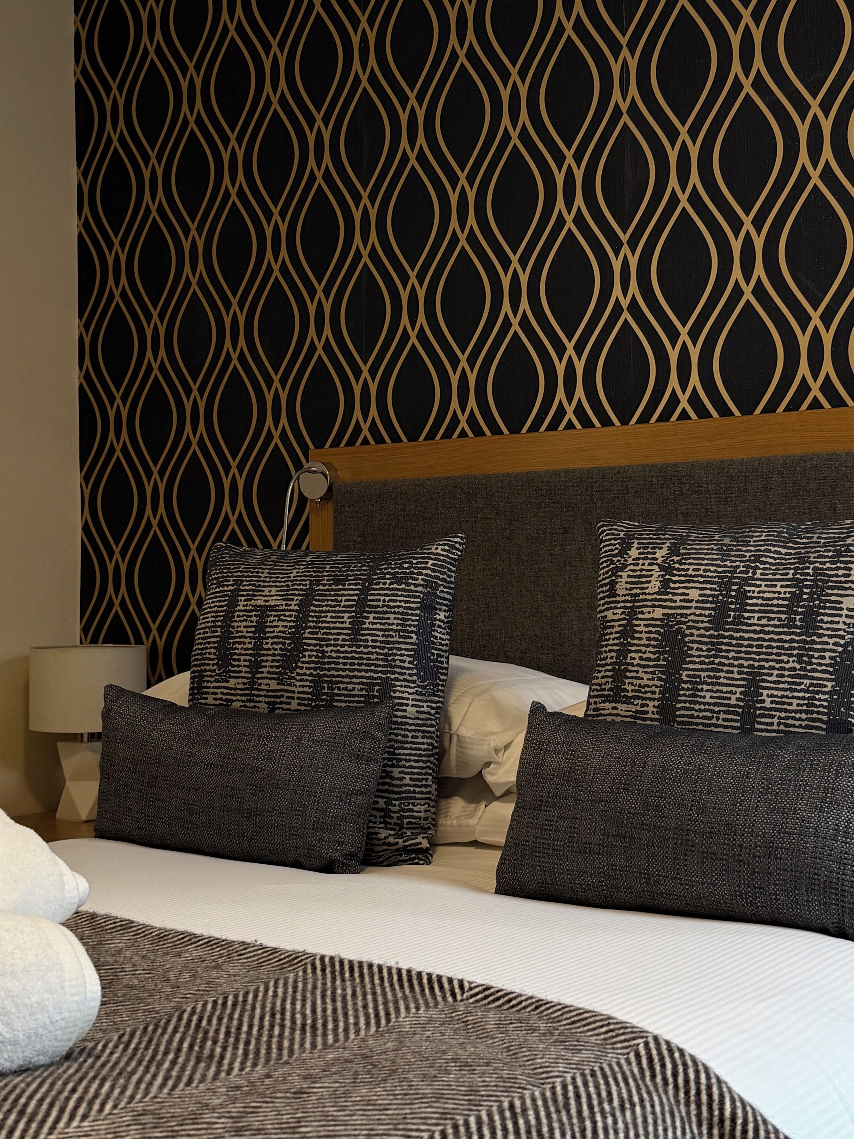 Een hotelkamer met een patroon op de muur, een bed met veel kussens en een bedlampje.