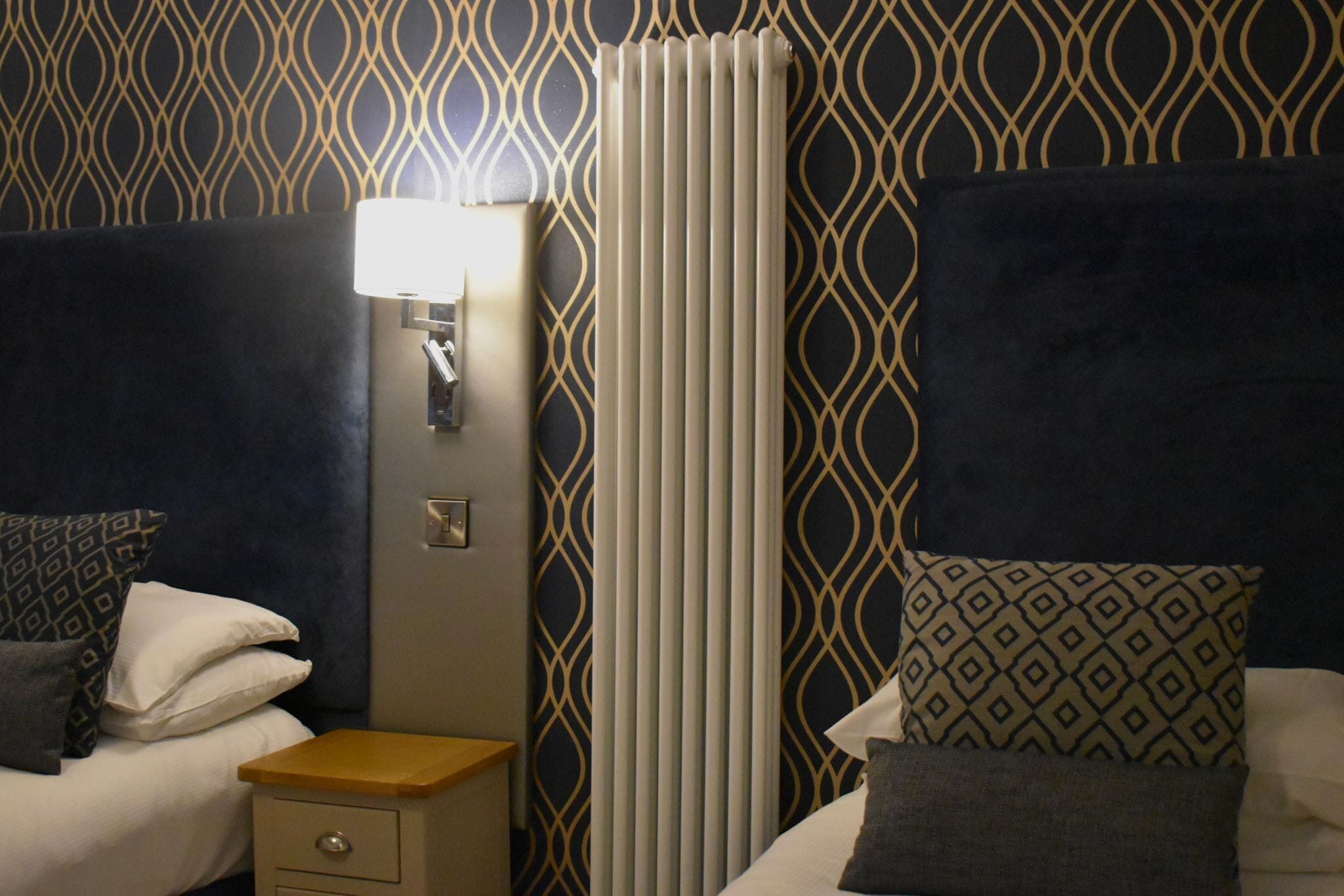 A hotel room with a patterned wallpaper, a bedside lamp, a nightstand, and a bed with pillows.