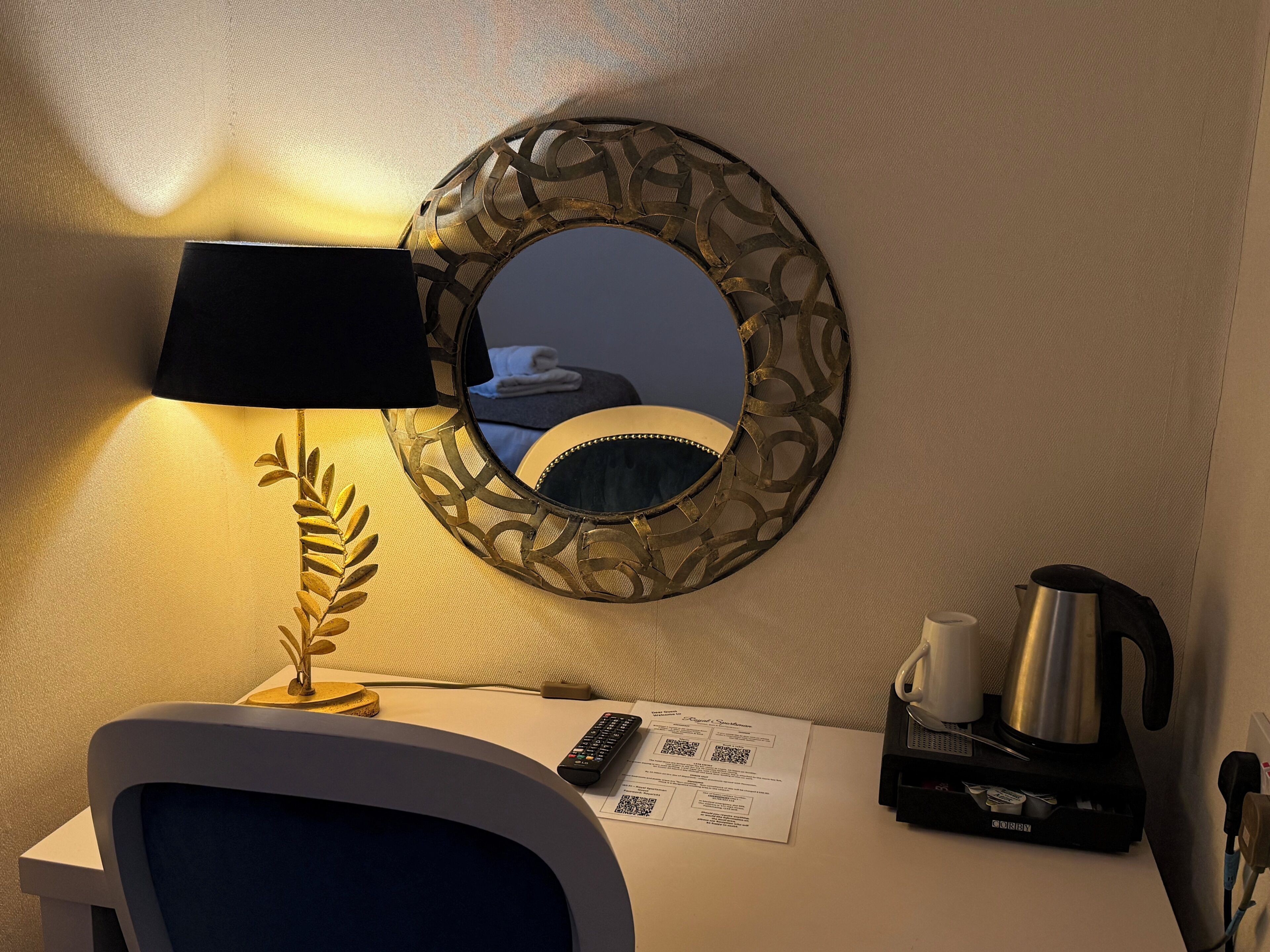 A hotel room with a desk, a lamp, a mirror, a remote control, a kettle, and a menu.