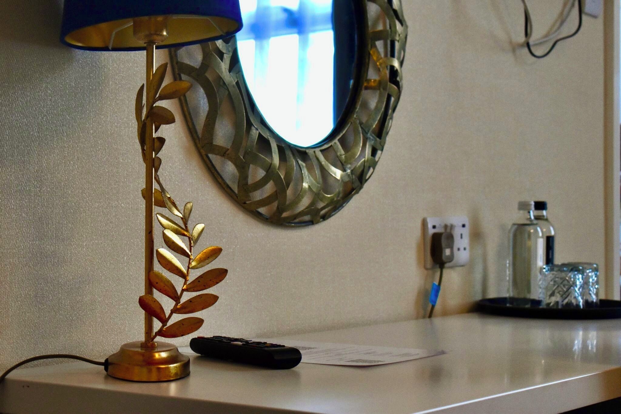 A desk with a lamp, a remote control, a mirror, a water bottle, and a power outlet.