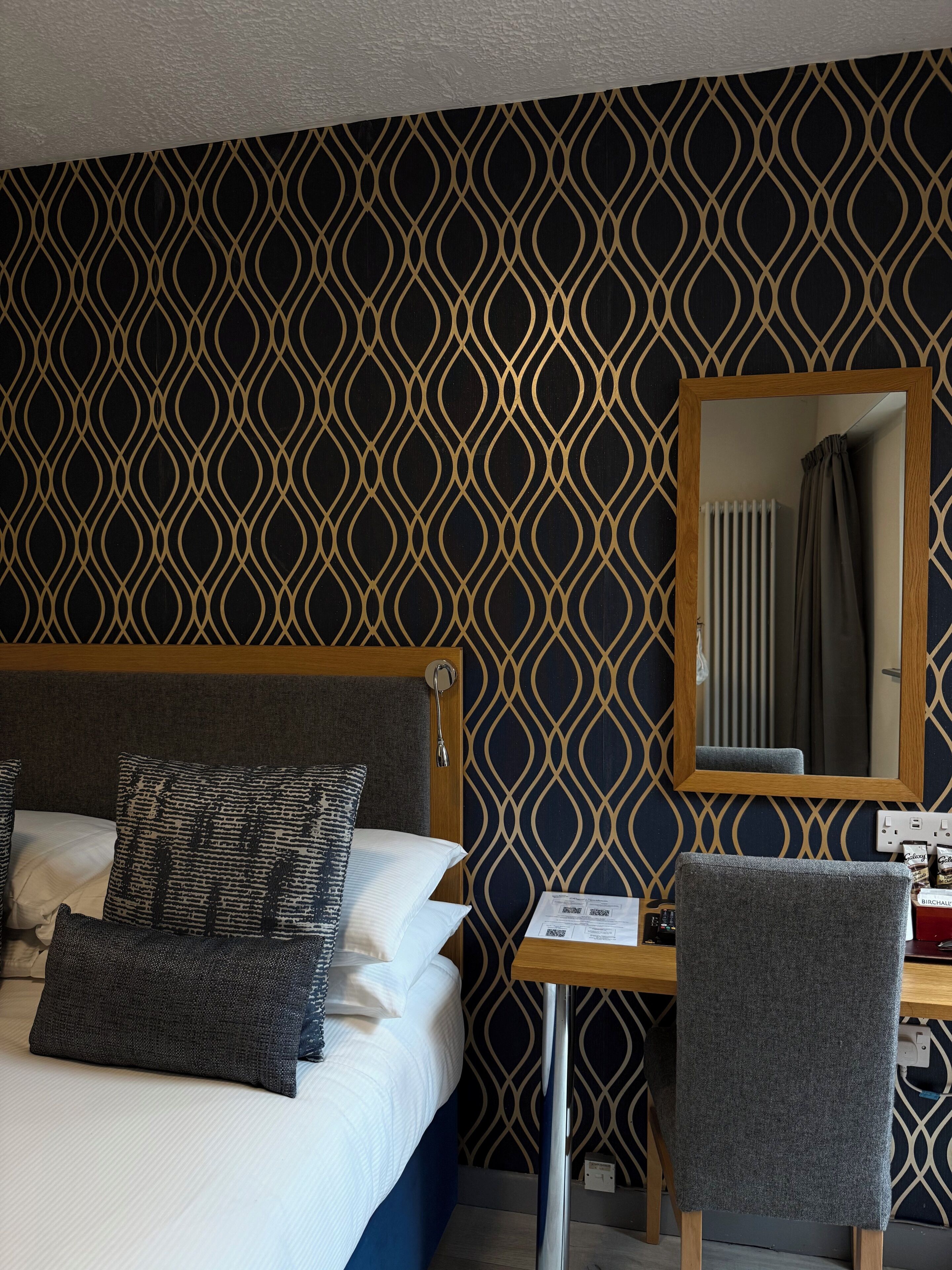 Une chambre d’hôtel avec un papier peint à motifs, un lit avec des oreillers, un bureau avec une chaise et un miroir.