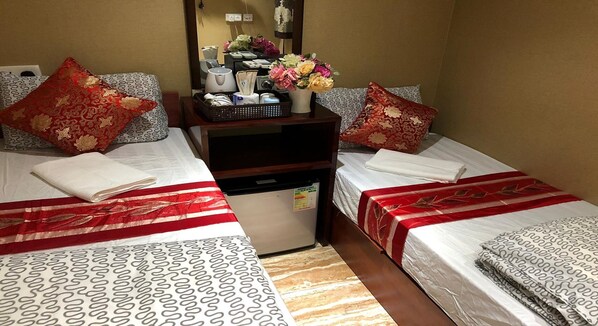 A hotel room with two beds, each with patterned bedding and red pillows. A small refrigerator and a desk with a mirror and a vase of flowers are in the center.