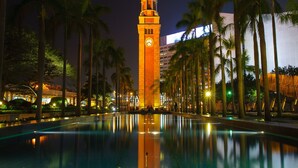 A nighttime view of a tall clock tower reflecting in a pool, surrounded by palm trees and modern buildings. - Rich Hostel (Kowloon)