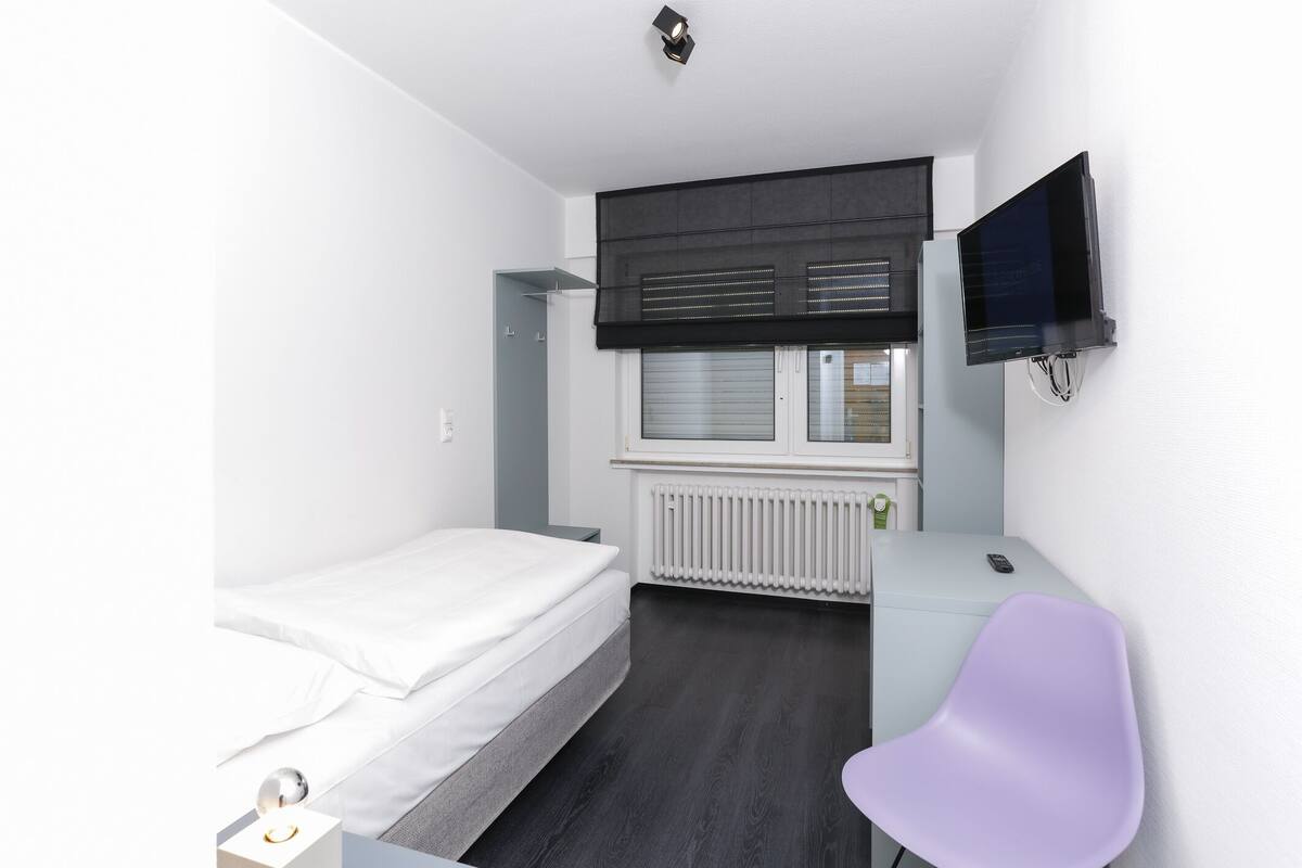 Un dormitorio moderno con una cama individual, un escritorio, una silla, un televisor y una ventana con persianas.