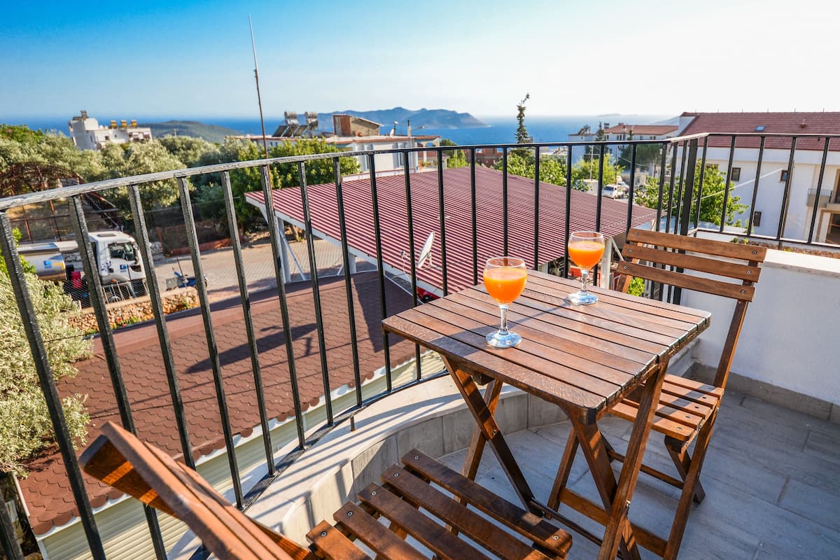 A balcony with a wooden table and chairs, a glass of orange juice, and a view of the sea and buildings.