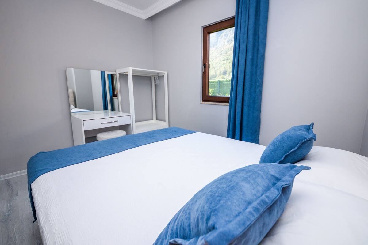 A bedroom with a bed, a bedside table, a mirror, and a window with curtains.