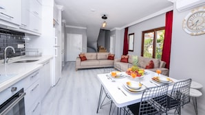 Una cocina moderna con armarios blancos, una zona de comedor con una mesa blanca y sillas negras, y un sofá con cojines rojos.