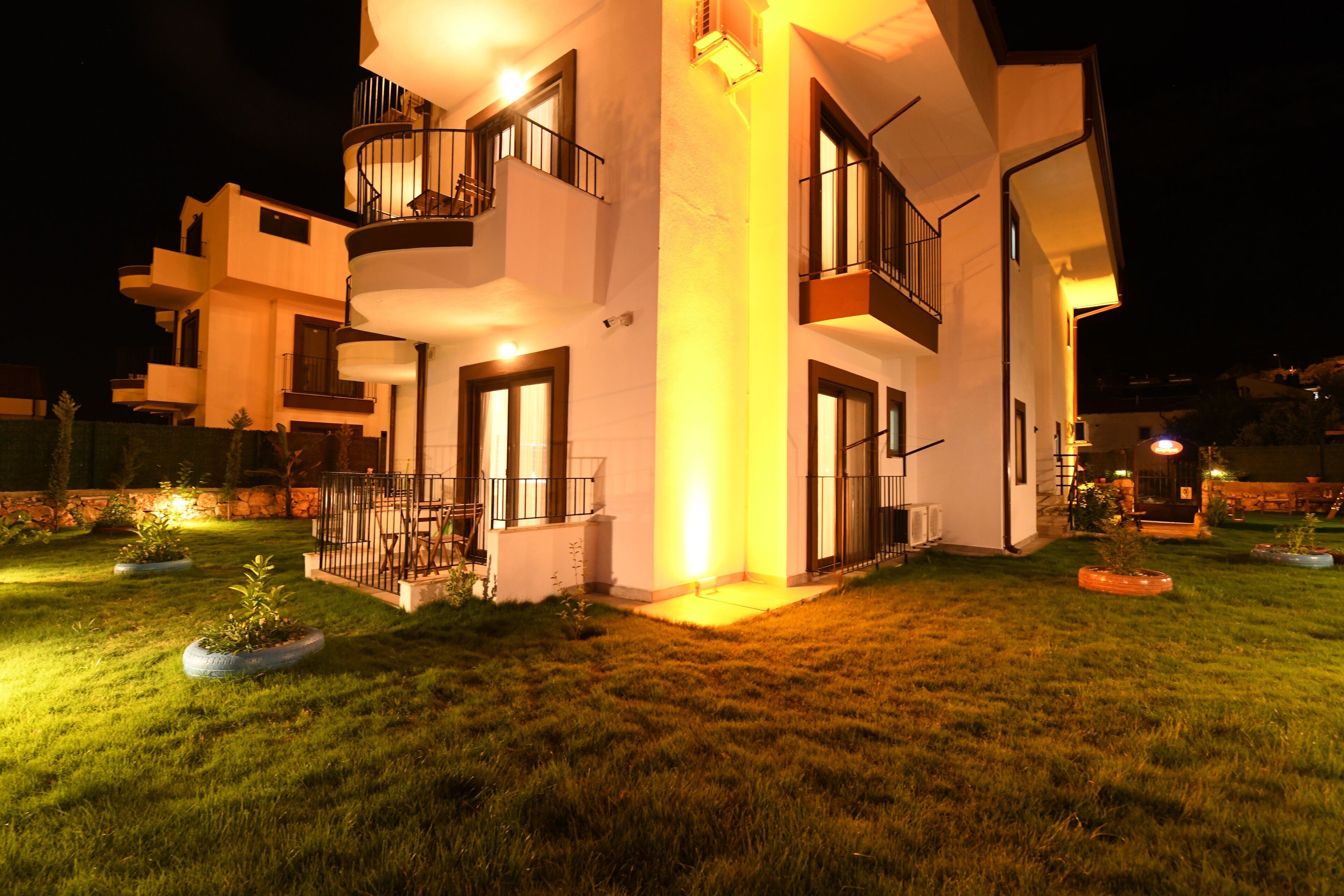 A modern two-story house with a balcony on the second floor, a well-lit entrance, and a grassy lawn.