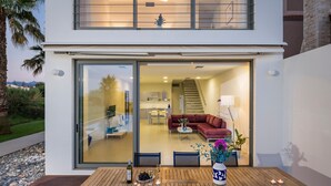 A modern house with a wooden deck, a dining area with a table and chairs, and a red couch inside.