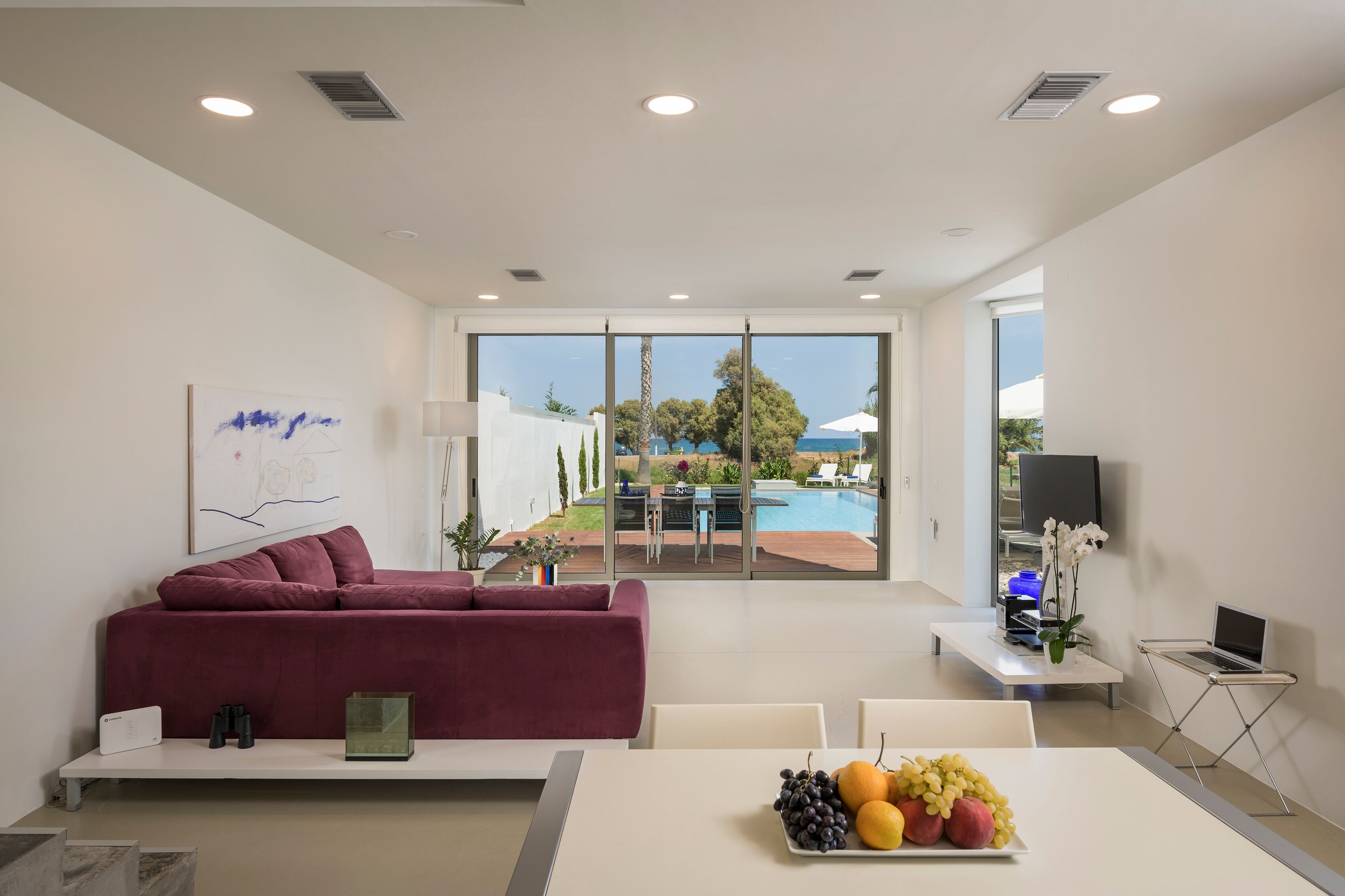 A modern living room with a sofa, a dining table with fruit, a TV, and a view of a pool and garden.