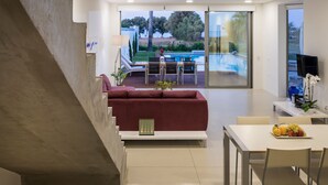 A modern interior with a staircase, a dining area with a table and chairs, a red sofa, and a view of a swimming pool and greenery outside.