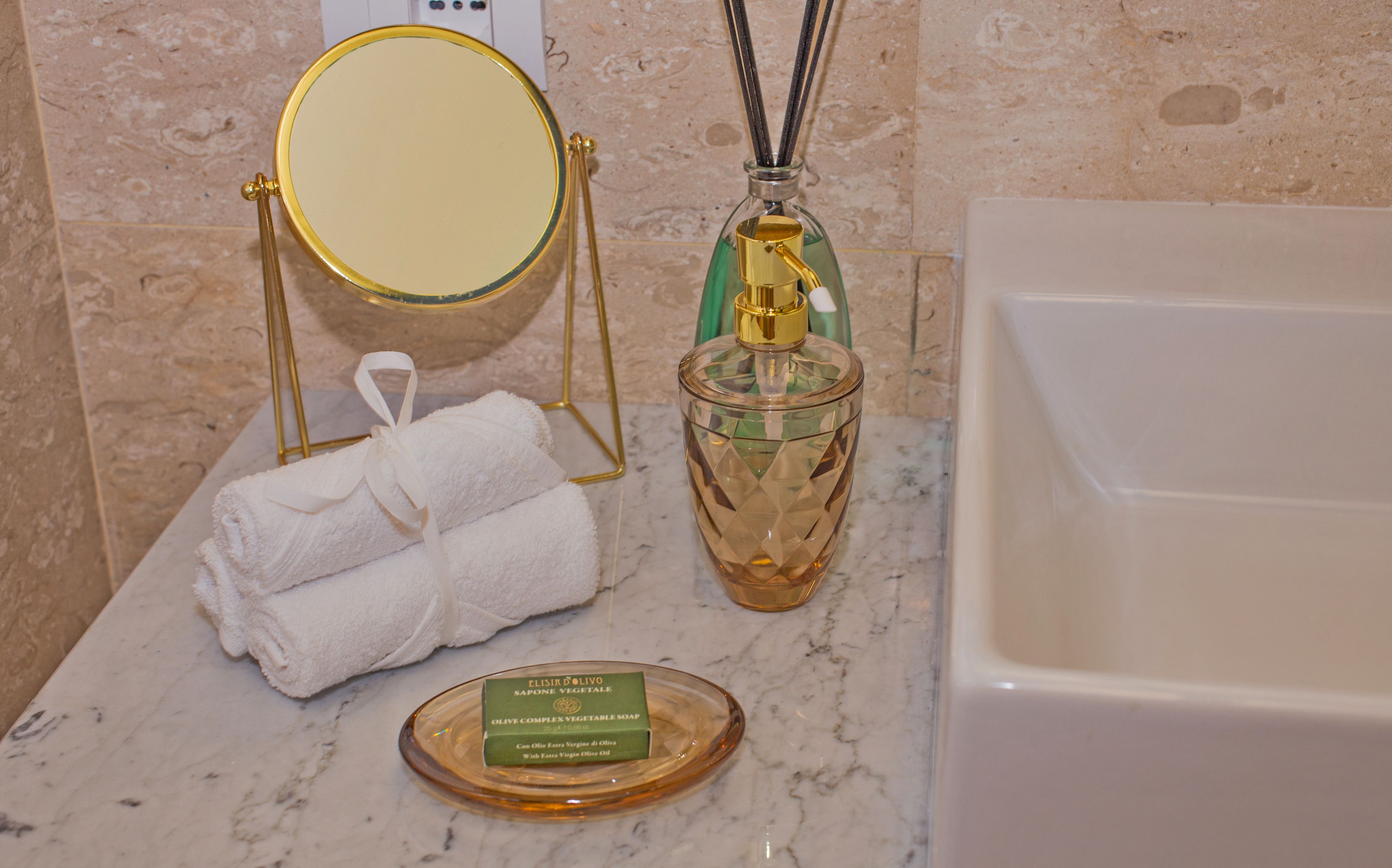 Een badkamer met een marmeren aanrechtblad, een spiegel met gouden lijst, een groene flesje rietverspreider, een witte handdoek en een fles olijfoliezeep.