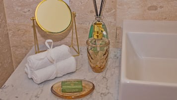 Een badkamer met een marmeren aanrechtblad, een spiegel met gouden lijst, een groene flesje rietverspreider, een witte handdoek en een fles olijfoliezeep.