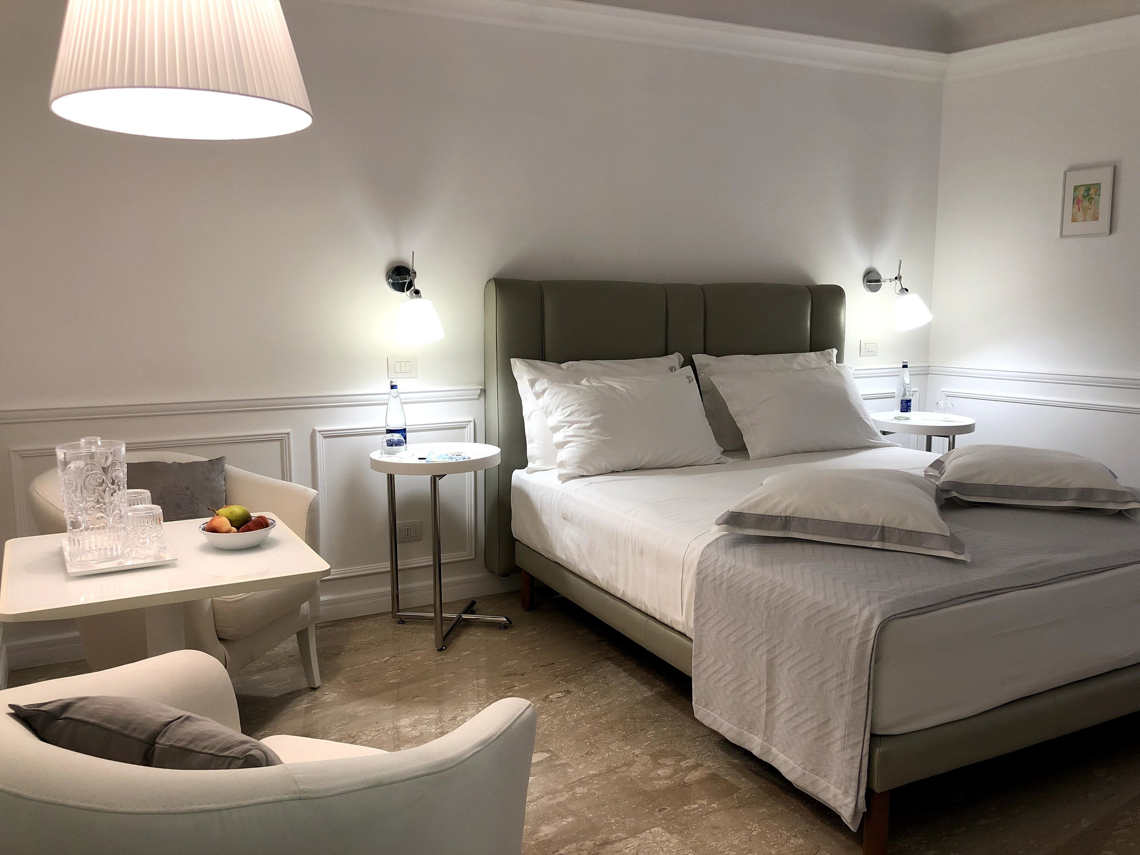 Una habitación de hotel con cama, mesitas de noche, una silla y una pequeña mesa con un frutero y una botella.
