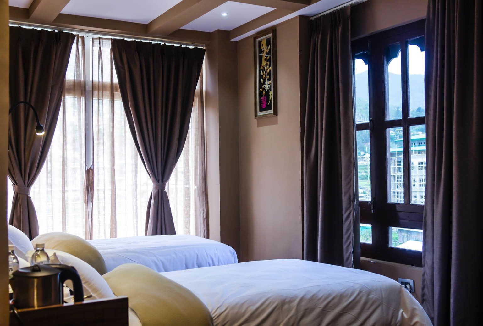 A hotel room with two beds, a large window with curtains, a painting on the wall, and a view of buildings outside.