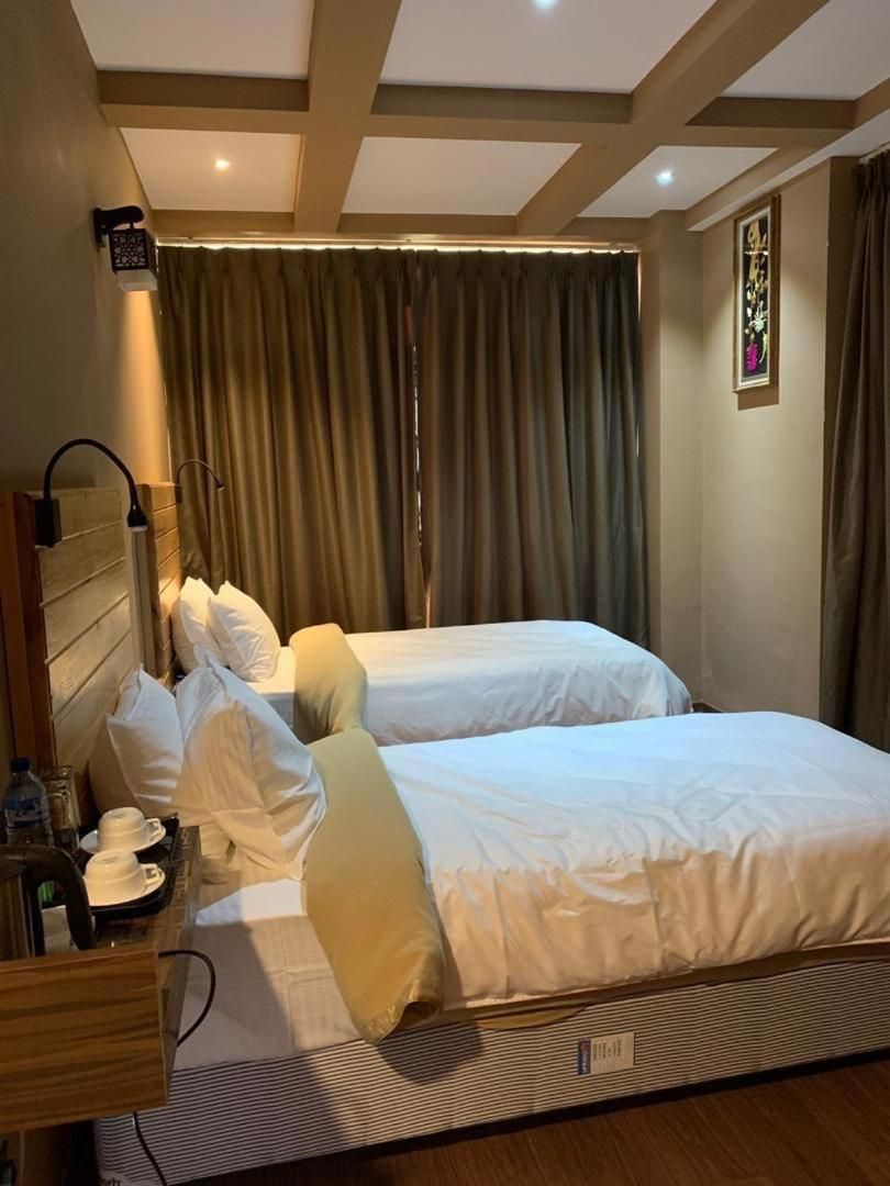 Habitación de hotel con dos camas, una mesita de noche con lámpara, una pequeña mesa con tazas y un teléfono, y un ventanal con cortinas.