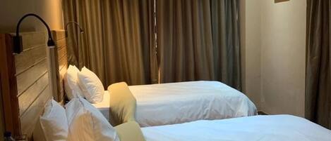Habitación de hotel con dos camas, una mesita de noche con lámpara, una pequeña mesa con tazas y un teléfono, y un ventanal con cortinas.