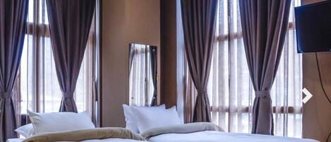 A hotel room with two beds, large windows with curtains, a flat-screen TV, and a mirror on the wall.